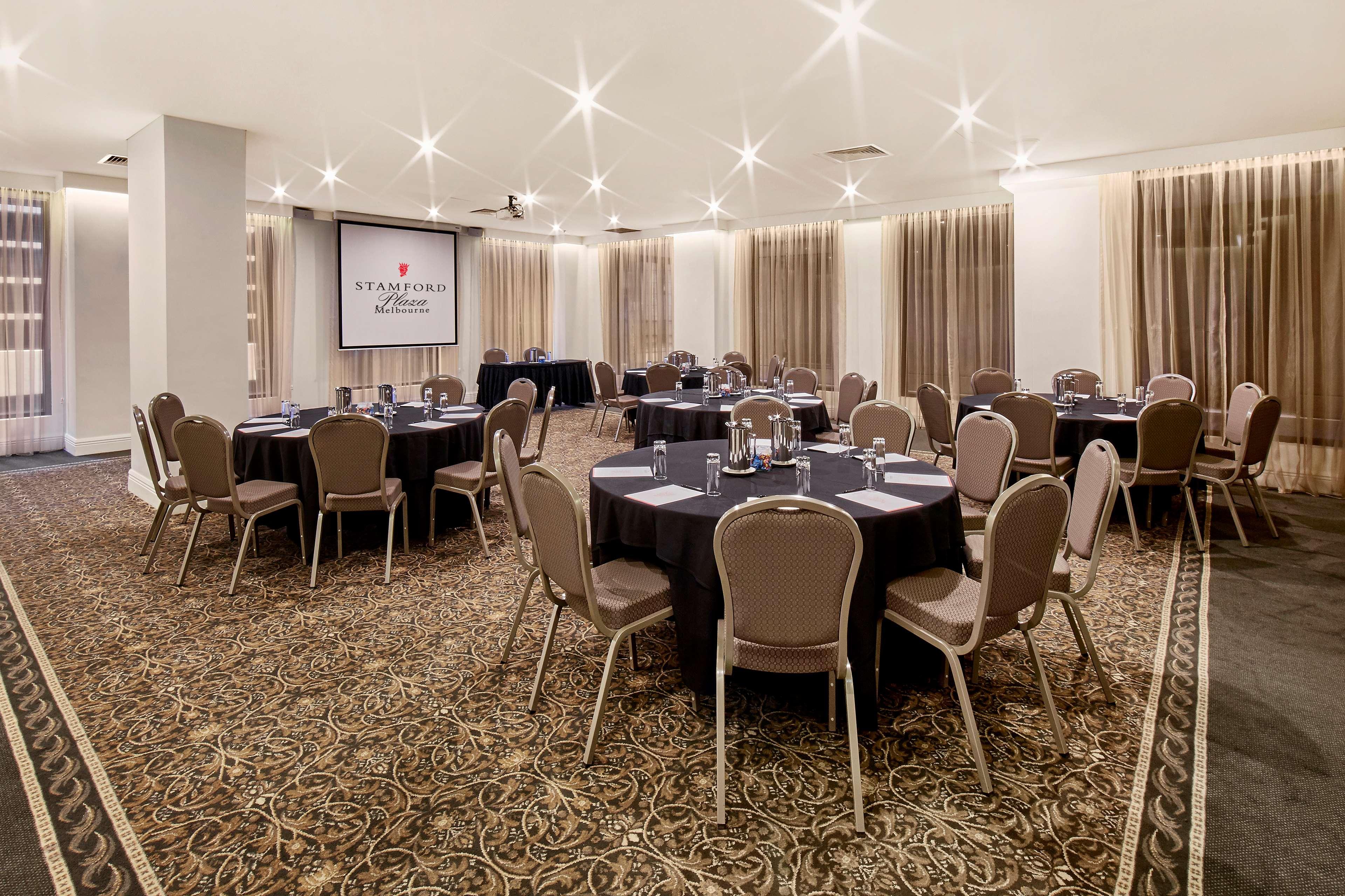 Hotel Stamford Plaza Melbourne City Exterior foto The photo shows a well-lit event space set up for a gathering or meeting. There are several round tables covered with black tablecloths, each surrounded by beige chairs. In the background, a large screen displays a logo or branding, likely related to