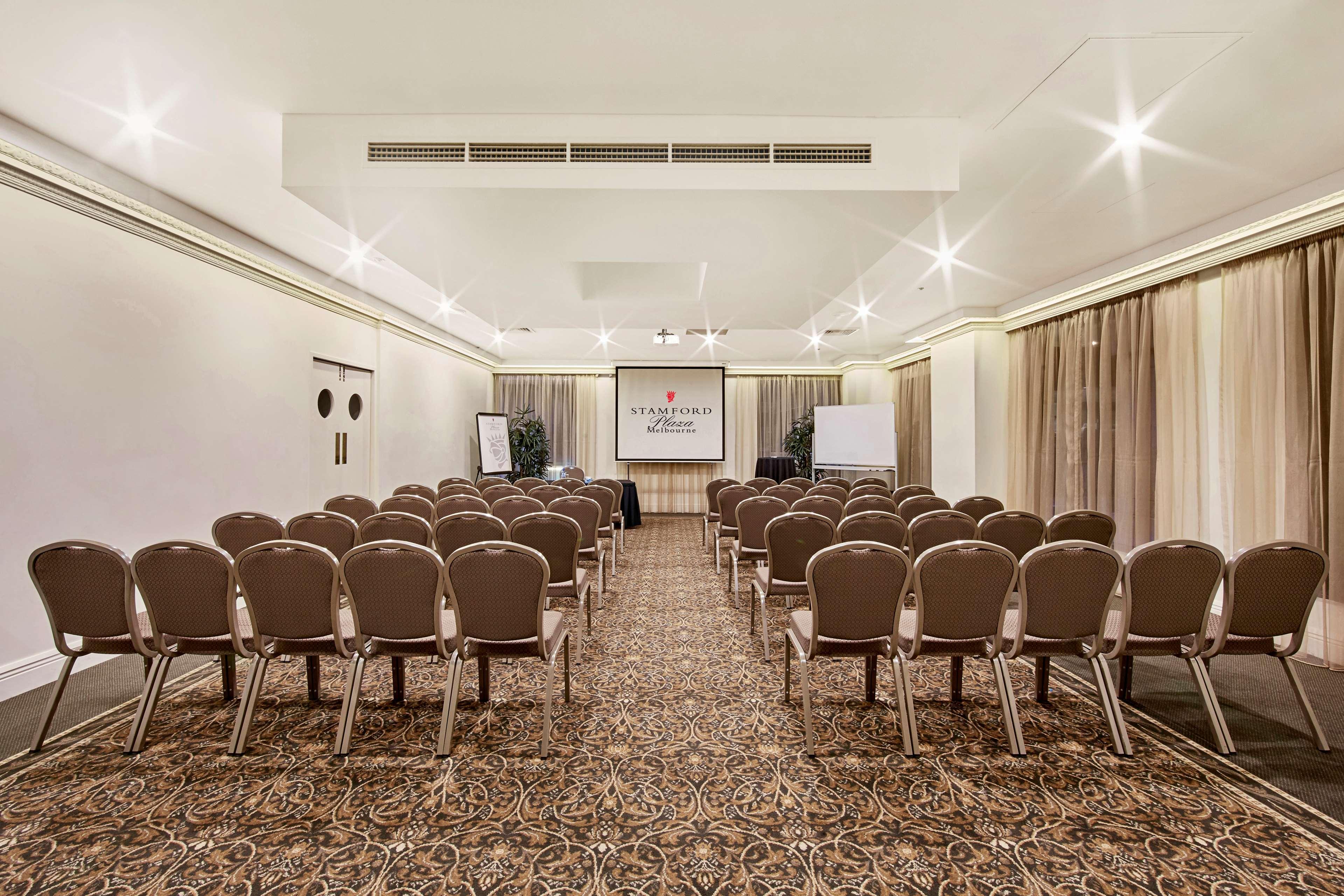 Hotel Stamford Plaza Melbourne City Exterior foto The photo shows a conference room set up for an event. It features several rows of beige chairs arranged facing a central presentation area. At the front, there is a screen displaying a logo or title, possibly for a conference or meeting. The walls h