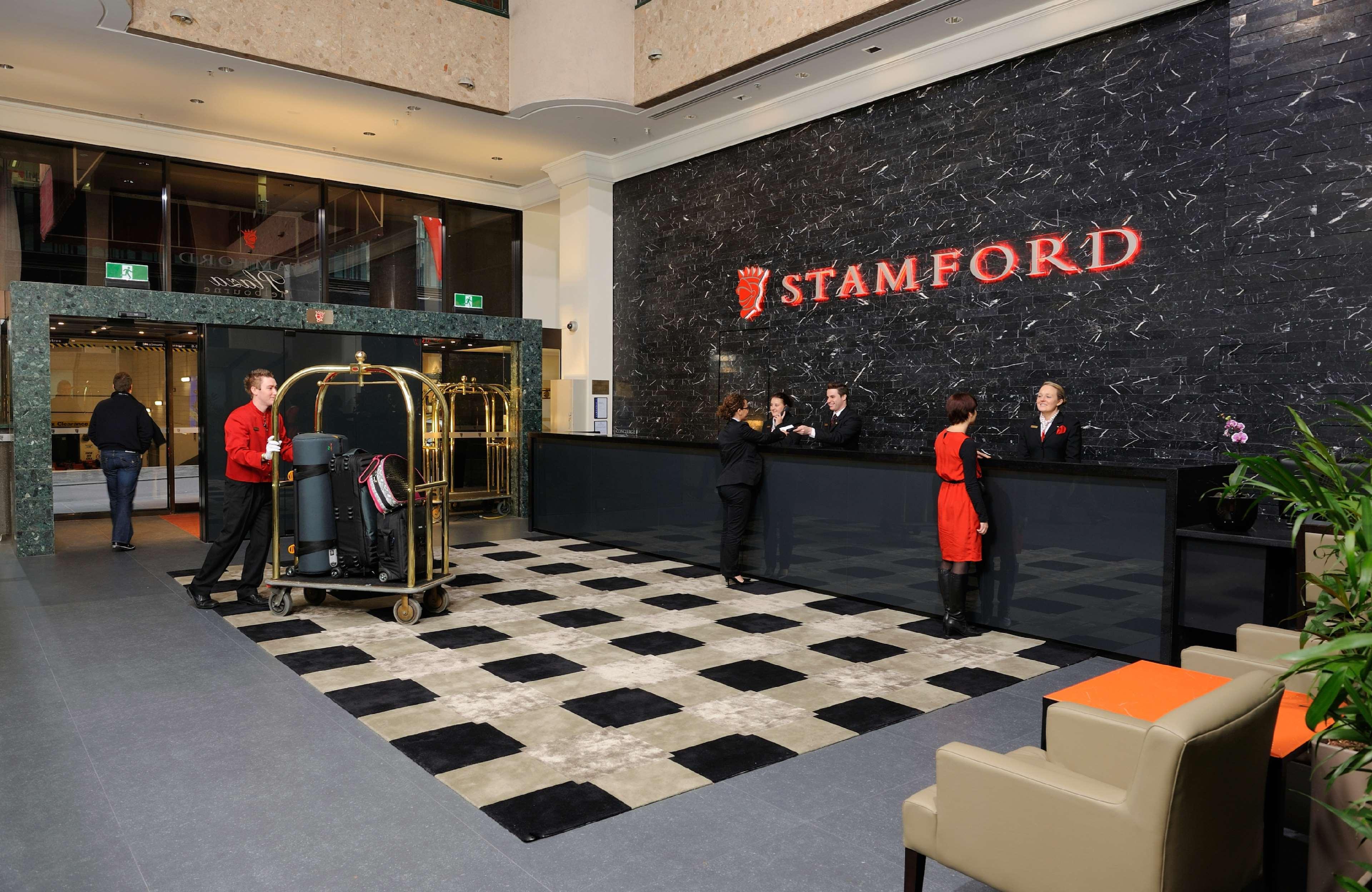Hotel Stamford Plaza Melbourne City Interior foto The photo shows the interior of a hotel lobby, specifically the Stamford Hotel. There is a large reception desk where several staff members are attending to guests. In the foreground, a bell cart loaded with luggage is positioned beside the reception