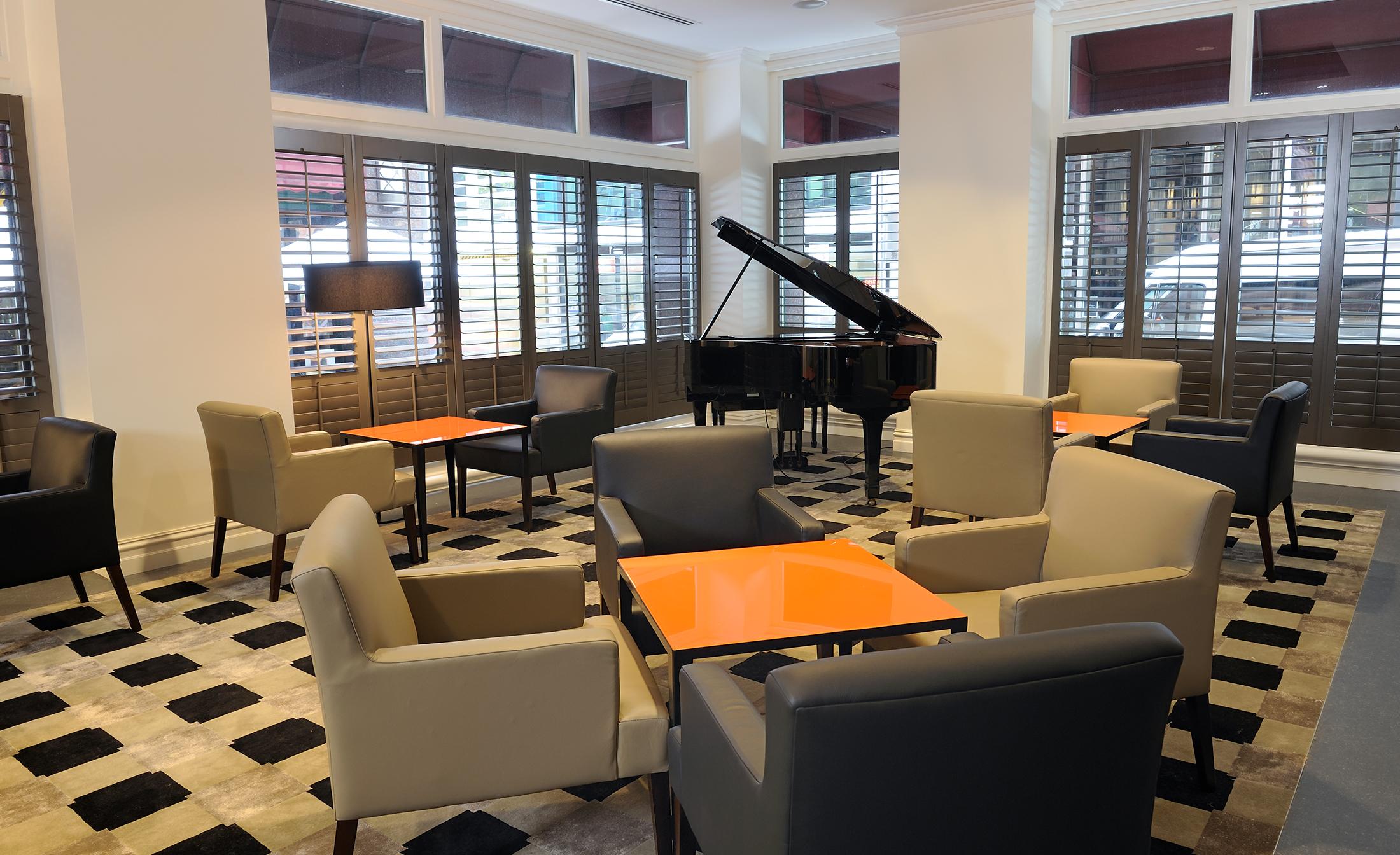 Hotel Stamford Plaza Melbourne City Exterior foto The photo shows a stylish indoor lounge area. It features several light gray armchairs arranged around orange square tables. In the corner, there is a black grand piano, adding an elegant touch to the space. The room has large windows with wooden shu