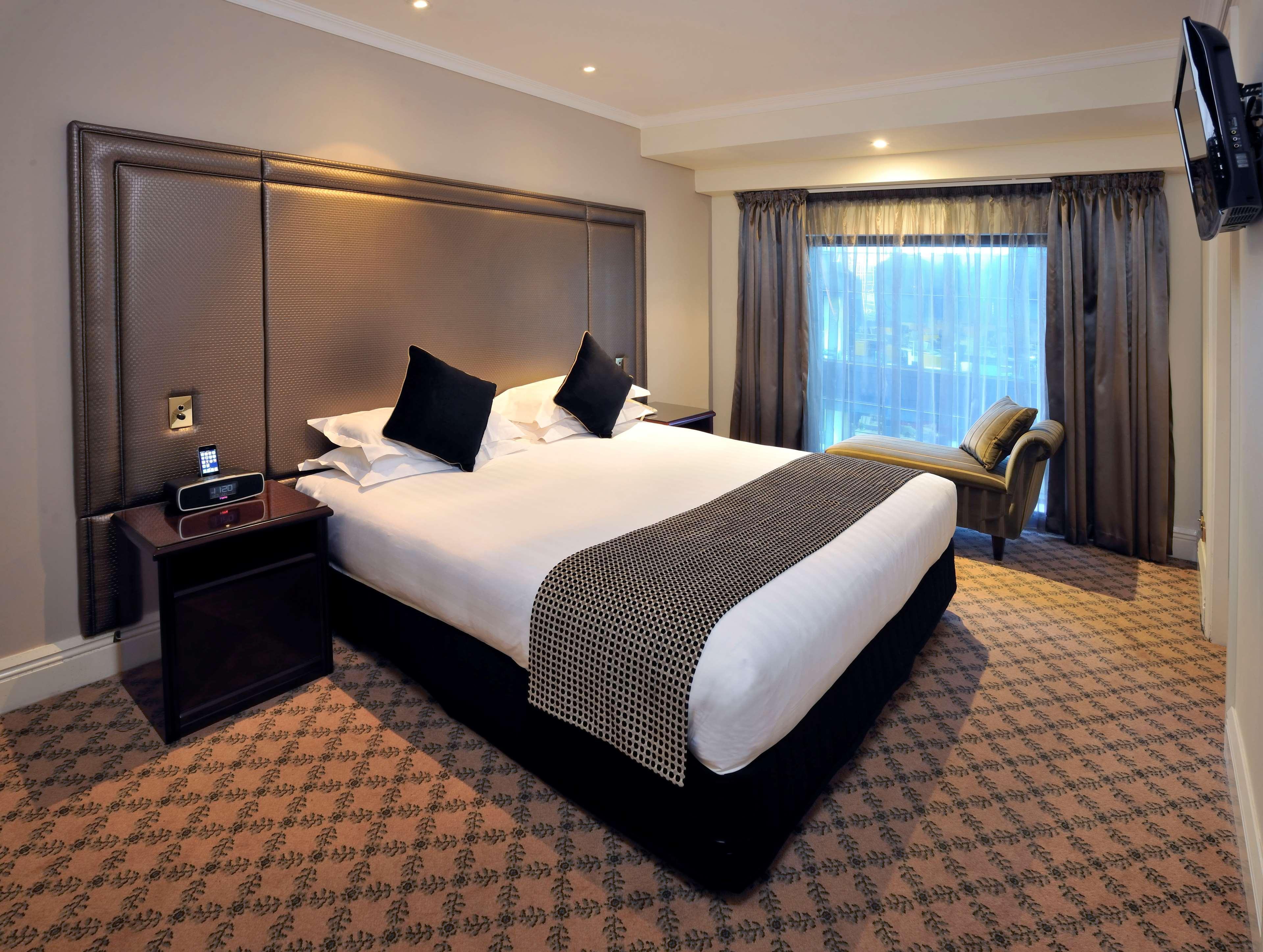 Hotel Stamford Plaza Melbourne City Exterior foto The photo shows a hotel room featuring a neatly made king-sized bed with dark pillows. The headboard is upholstered in a dark material, and there is a decorative blanket at the foot of the bed. Beside the bed are two nightstands, each equipped with l
