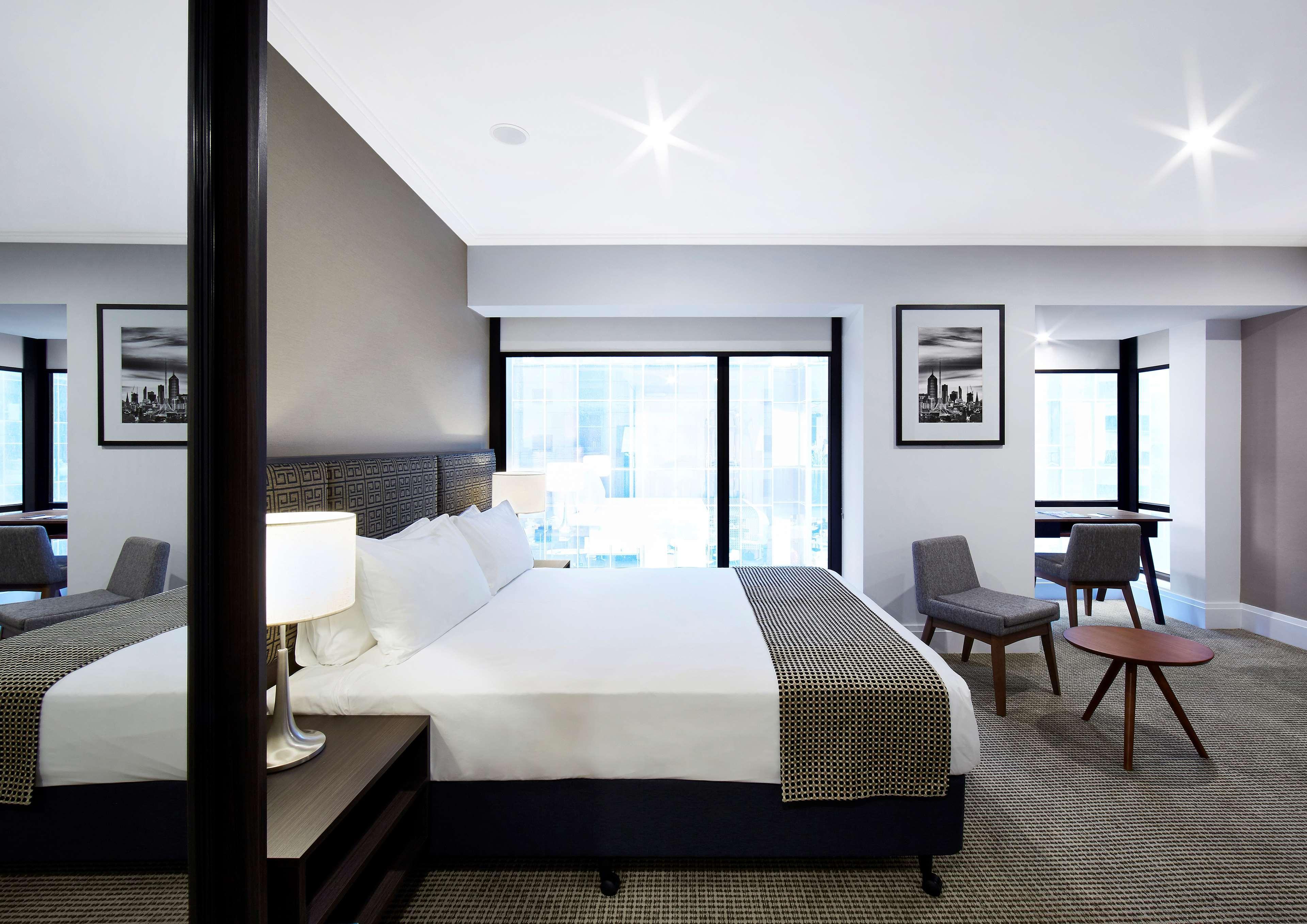 Hotel Stamford Plaza Melbourne City Exterior foto The image shows a modern hotel room featuring a large bed with white linens and a gray throw blanket. There are two windows that allow natural light to enter the space, and the walls are painted in a light color. In addition to the bed, there are two