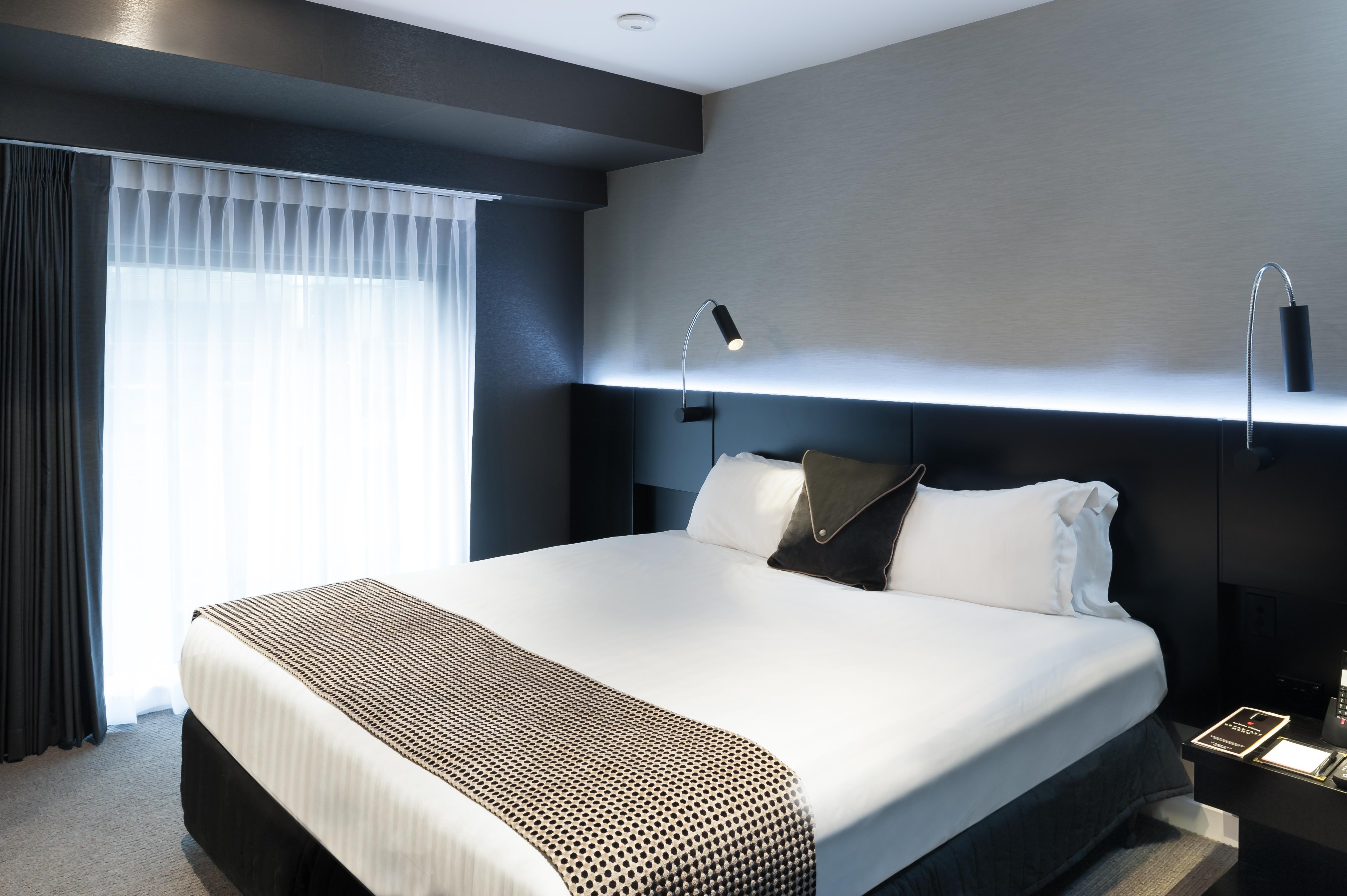 Hotel Stamford Plaza Melbourne City Exterior foto The photo shows a modern hotel room featuring a large bed with white linens and a decorative throw blanket. The room has a sleek and contemporary design, with gray walls and a soft lighting strip along the headboard. A window is adorned with sheer cu