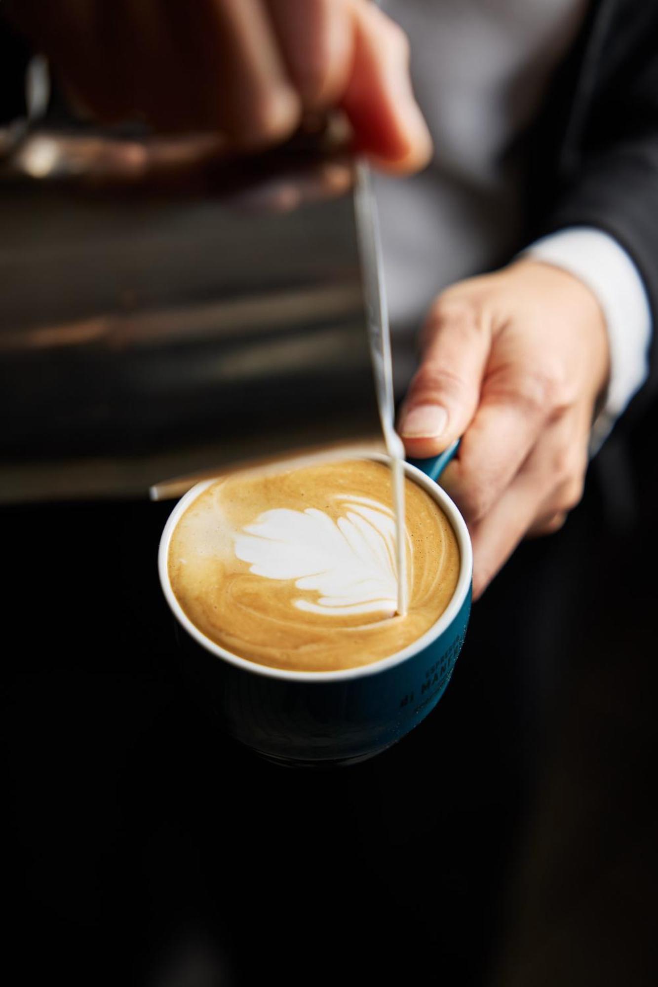 Hotel Stamford Plaza Melbourne City Exterior foto The photo shows a close-up of a hand holding a cup of coffee with a latte art design on the surface. The coffee appears to be a creamy, caramel color, likely a latte or cappuccino. A small stream of milk is being poured into the cup, creating a heart