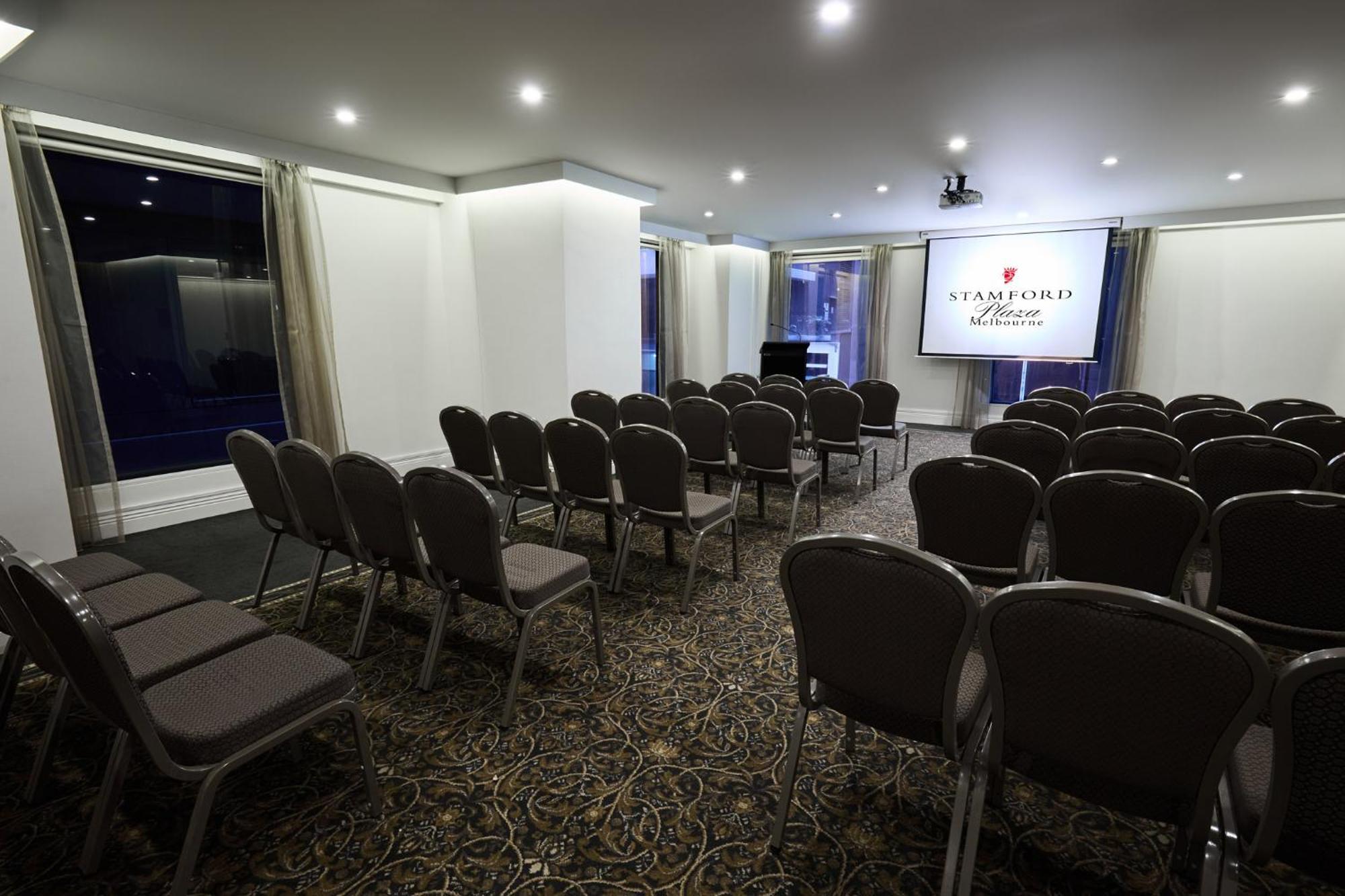Hotel Stamford Plaza Melbourne City Exterior foto The image shows a conference or meeting room set up for an event. There are rows of gray chairs arranged facing a large screen displaying the name "Warford House." The room is well-lit with overhead lighting and has large windows covered with light-c