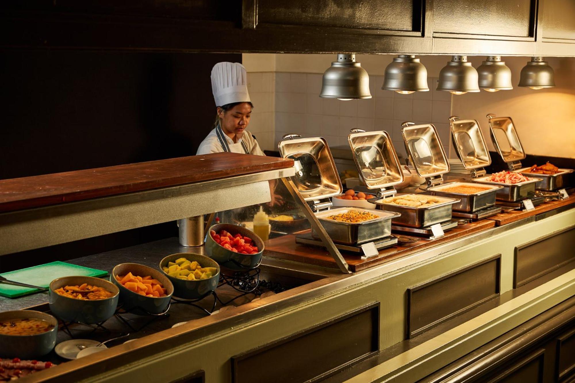 Hotel Stamford Plaza Melbourne City Exterior foto The photo shows a buffet setup in a dining area. There is a chef in a white uniform and hat, standing behind a counter filled with various food options. The buffet features several metal serving trays containing different dishes, with warming lights 