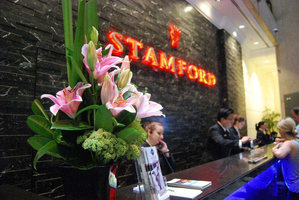 Hotel Stamford Plaza Melbourne City Exterior foto The photo shows a hotel reception area with a prominent sign that reads "STAMFORD" in bright red letters. In the foreground, there is a decorative arrangement of flowers, primarily featuring pink lilies, along with greenery. The reception counter is 